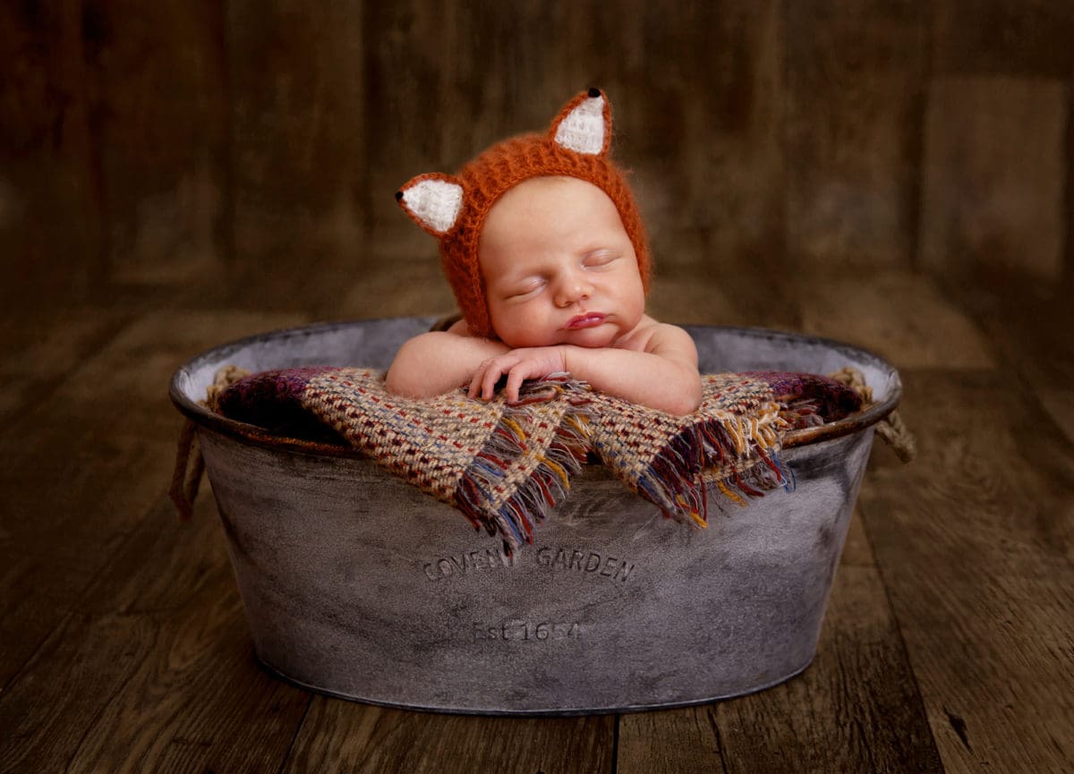 Photograph of sleeping newborn baby in cute crocheted fox outfit in an old tin bath